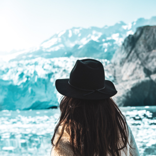Woman wearing a hat looking at an iceberg with a mountain in the background.
